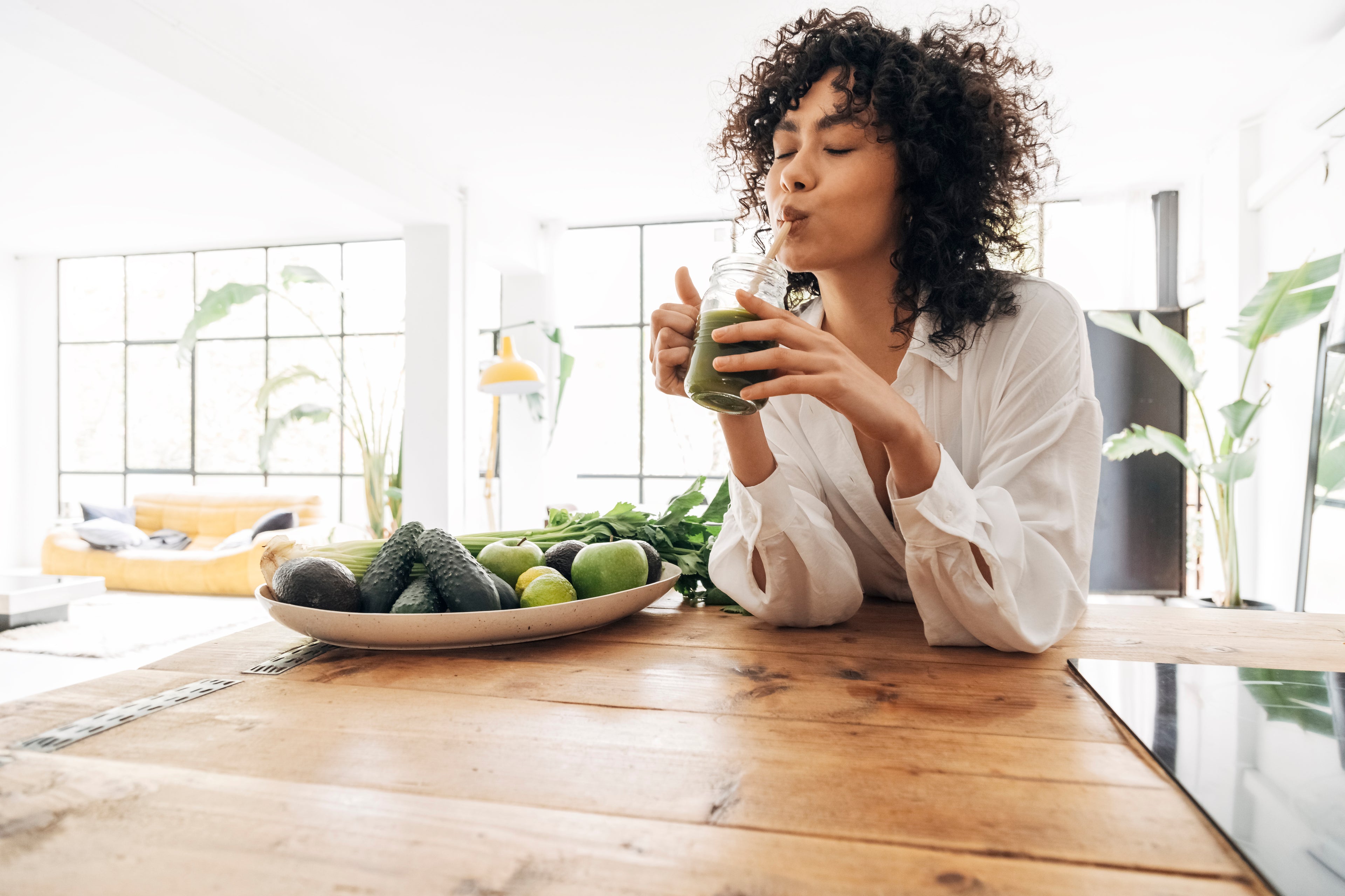 young lady with dark curly hair drinking green smoothie with fruit and vegetables next to her
